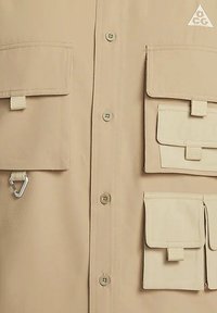 Beige skjorta med knappar som har flera fickor, inklusive fyra klaffar, en karbinhake och en triangulär logotyp i det övre högra hörnet.