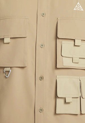 Beige kauluspaita, jossa on useita taskuja, mukaan lukien neljä läppätaskua, karabiinihaarniska ja kolmiomainen logo oikeassa yläkulmassa.