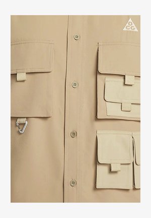 Chemise beige à boutons avec plusieurs poches, dont quatre à rabat, un point d'attache pour un mousqueton et un logo triangulaire dans le coin supérieur droit.