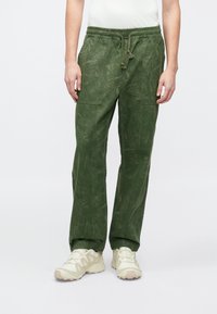 Pantalones cargo verdes con un acabado texturizado, que cuentan con una cinturilla elástica y dos grandes bolsillos delanteros. Combinados con calzado de color claro.