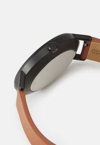 Svart metallkapsel med cirkulär form, brun läderrem och en tryckknapp på sidan. Slät, matt yta på baksidan.