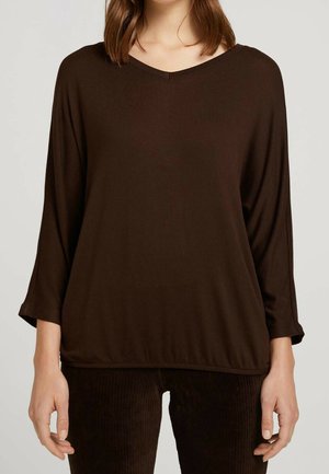 Frau trägt ein locker sitzendes, langärmliges, dunkelbraunes V-Ausschnitt-Oberteil mit passender dunkelbrauner, strukturierter Hose vor einfachem Hintergrund.