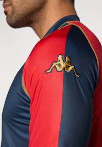 Długi rękaw koszulki sportowej z czerwonymi i granatowymi panelami, z wyszywanym na złoto logo dwóch postaci na ramieniu. Gładka, elastyczna tkanina.