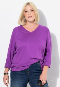 Lila V-Ausschnitt-Shirt mit langen Ärmeln, hochgerollten Bündchen und lockerer Passform, kombiniert mit dunkler Jeans. Glatter Stoff ohne sichtbare Muster.