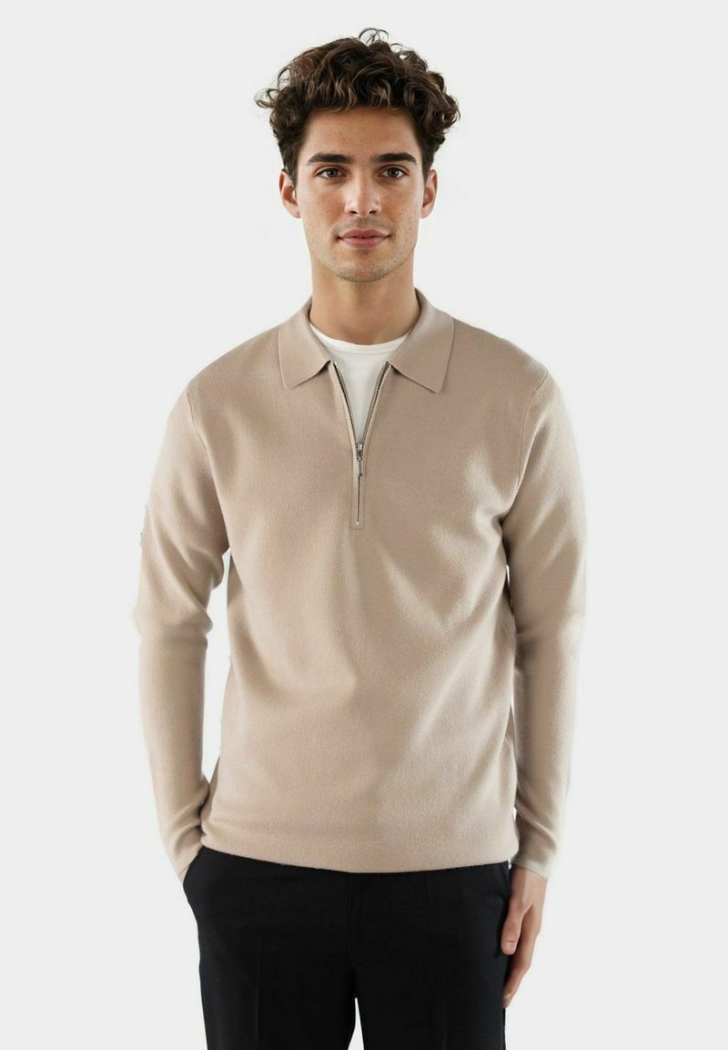 Beige Pullover mit Reißverschlusskragen, langen Ärmeln und weicher Textur. Über einem weißen Hemd getragen, kombiniert mit dunklen Hosen.