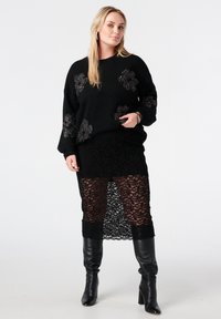 Pull en maille noire avec motifs floraux et tissu texturé, associé à une jupe en dentelle noire transparente et des bottes en cuir noires montantes jusqu'au genou.