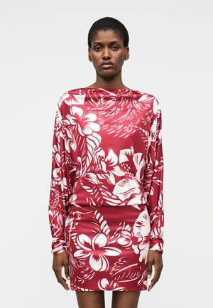 Γυναίκα που φοράει κόκκινο φόρεμα με μακριά μανίκια με μεγάλα λευκά λουλουδένια και φυλλώδη σχέδια, στέκεται μπροστά σε απλή ανοιχτόχρωμη επιφάνεια.