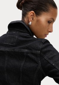 Svart denimjacka med hög krage, grå sömmar och detaljer i ryggen. Har en slät, mjuk textur och en figursydd design.