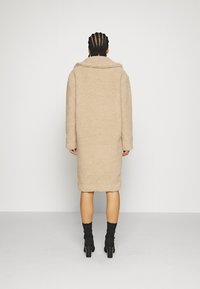 Beiger, übergroßer Mantel mit strukturierter Oberfläche, ausgestattet mit einem breiten Kragen und langen Ärmeln, die bis zur Mitte der Wade reichen. Kombiniert mit schwarzen Knöchelsocken und High Heels.