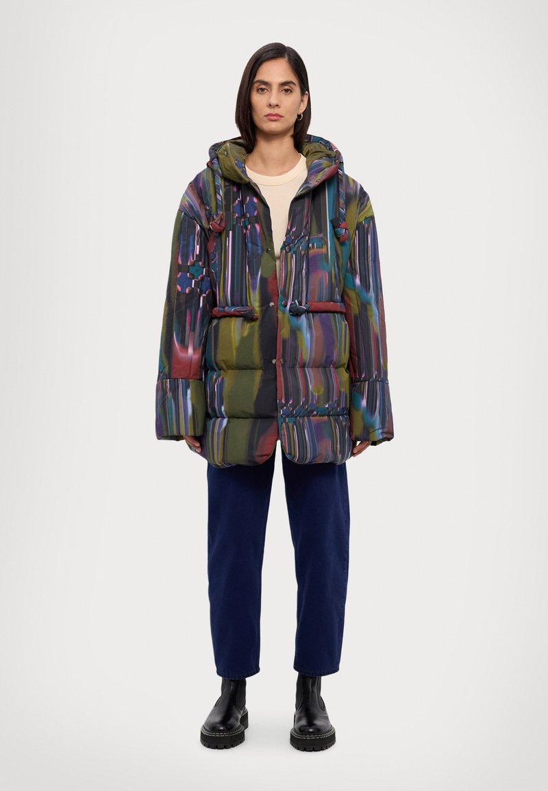 Doudoune avec un motif multicolore, dotée d'une capuche, d'accents à cordon de serrage et de deux poches avant. Portée avec un pantalon bleu foncé et des bottes noires.