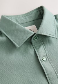 Gros plan sur le col d'un polo vert clair texturé et deux boutons, montrant le tissage du tissu et les détails des coutures.