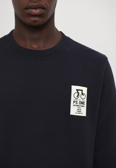 Laivansininen collegepaita, joka on valmistettu puuvillasta ja jossa on pyöreä kaula-aukko sekä suorakulmainen valkoinen läiskä, jossa on polkupyöräkuva ja useita kaupunkinimiä.