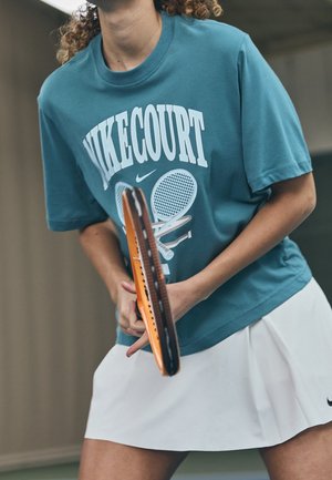 Person hält einen orangefarbenen Tennisschläger, trägt ein türkisfarbenes "NIKECOURT"-Shirt und einen weißen Rock und bereitet sich darauf vor, in der Halle zu spielen.