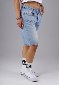 Hellblaue Denim-Bermuda-Shorts mit lockerer Passform, fünf Taschen, Metallbeschlägen und einem rohen Saum. Kombiniert mit weißen High-Top-Sneakern.
