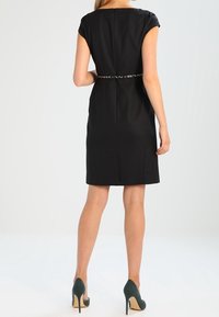 Sort kjole med korte ærmer, tætsiddende silhuet og et dekorativt bælte. Glat stof, knælængde, kombineret med grønne høje hæle.