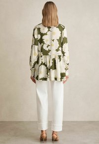 Top peplum floreale verde con grandi fiori bianchi, abbinato a pantaloni bianchi e scarpe con tacco marroni. Tessuto morbido, maniche lunghe, vestibilità casual.