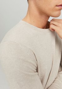 Homme portant un pull beige clair, main ajustant le col près du cou, fond neutre.