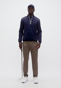 Άνδρας με μπλε πουλόβερ με φερμουάρ μέχρι τη μέση, μπεζ παντελόνι, λευκά αθλητικά παπούτσια και καπέλο, κρατώντας ένα μπαστούνι του γκολφ και στέκεται μπροστά σε απλό φόντο.