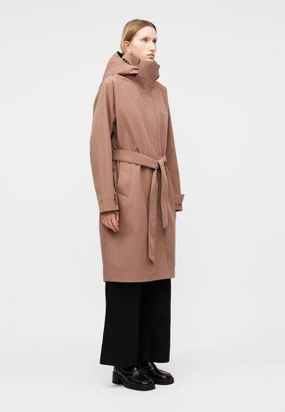 Žena stojaca v hnedom kabáte po kolená s opaskom a kapucňou, čiernych širokých nohaviciach a čiernych topánkach na bielom pozadí.