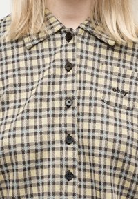 Κοντινό πλάνο ενός πουκαμίσου με κίτρινους και μαύρους καρό, με τσέπη που έχει κεντημένη τη λέξη "obey".