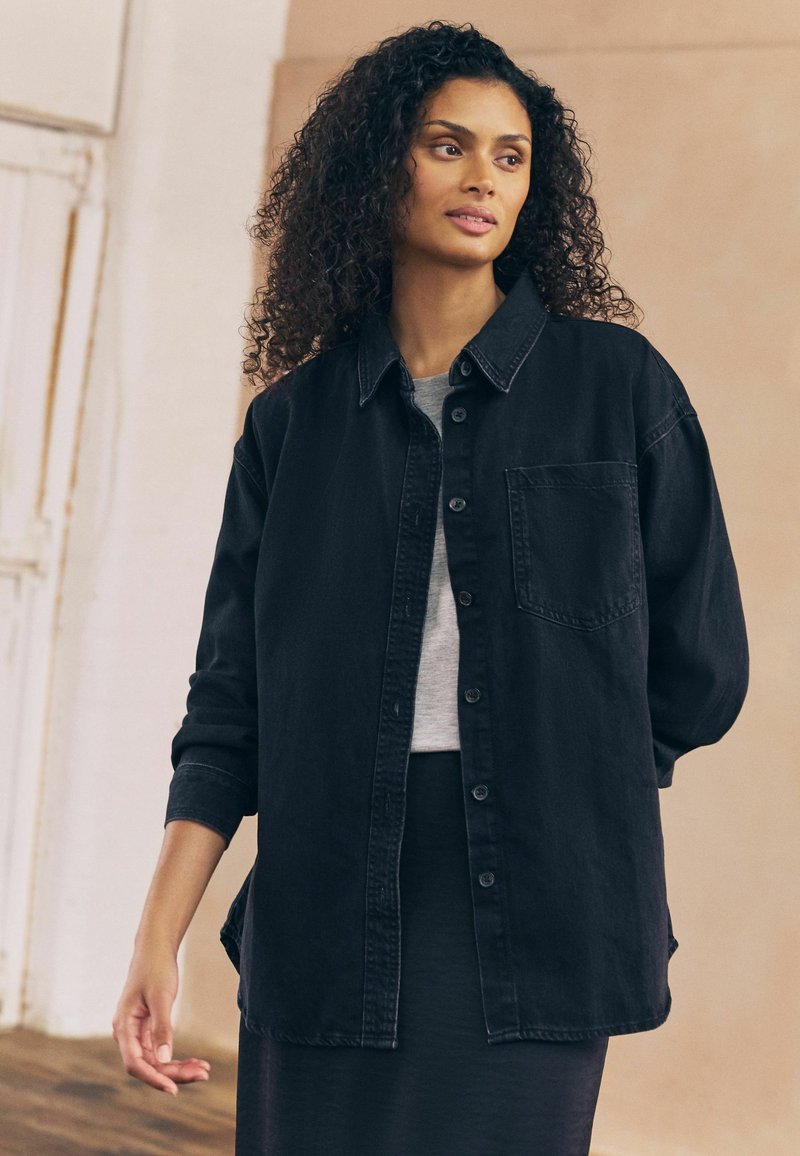 Zwarte denim overhemdjas met een relaxte pasvorm, knoopsluiting en zak; gecombineerd met een lichtgrijs topje en een donkere rok.