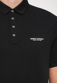 Polo noir avec quatre boutons et logo blanc Armani Exchange sur la poitrine gauche indiquant Milano New York.