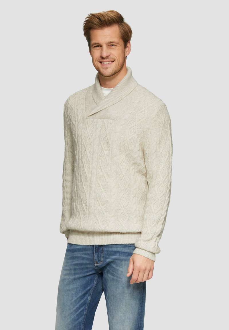 Helle beige Strickjacke mit Schalkragen, ausgestattet mit einem Rautenmuster und gerippten Bündchen. Kombiniert mit blauen Denim-Jeans.