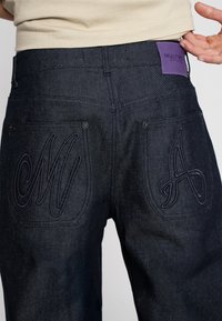 Jeans en denim foncé avec une texture subtile, patch de marque violet à la taille et grande broderie "M" et "A" sur les poches arrière.
