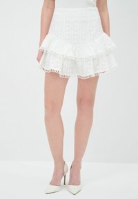 Jupe tiered blanche en tissu texturé avec des bordures en dentelle. Comprend trois couches froncées et une taille ajustée. Associée à des talons hauts blancs.