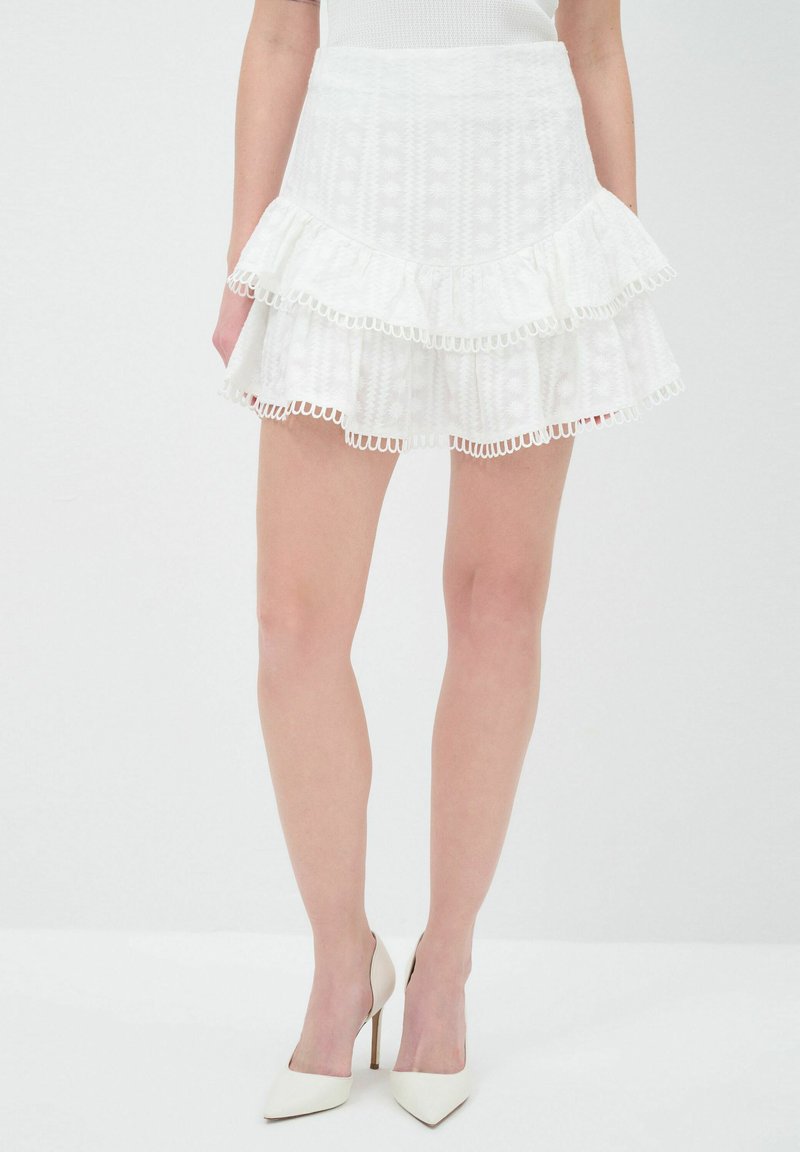Jupe tiered blanche en tissu texturé avec des bordures en dentelle. Comprend trois couches froncées et une taille ajustée. Associée à des talons hauts blancs.