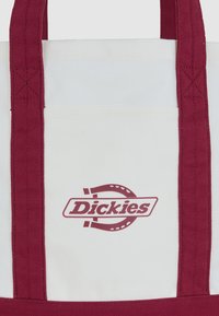 Biała torba płócienna z bordowymi uchwytami i bordowym logo "Dickies" z motywem podkowy na przedniej kieszeni.