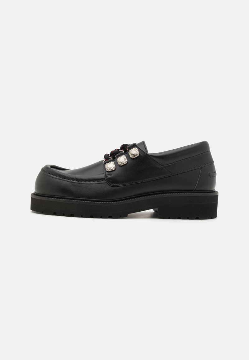 Mocasines de cuero negro con punta redonda, que presentan tres acentos metálicos y una suela texturizada para mayor agarre. Parte superior lisa y detalle de costura.
