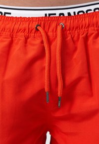 Shorts de bain rouges avec une taille élastique, ornés d'un détail logo noir et blanc et de cordons de serrage orange avec des embouts en argent. Texture lisse.