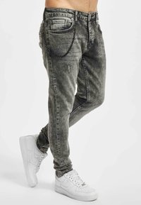 Jeans de denim griș, slim-fit, cu o textură ștearsă, dotate cu cinci buzunare și un accent de lanț negru pe buzunarul din spate. Asortate cu teniși albi.