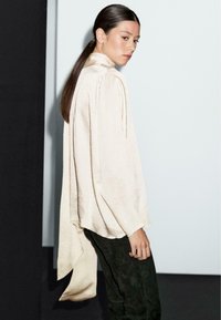 Blusa di colore crema, con texture, colletto alto e maniche fluide e drappeggiate. Abbinata a pantaloni verde scuro. Design semplice ed elegante.