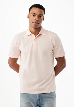 Mann steht mit den Händen hinter dem Rücken, trägt ein hellrosa kurzärmliges Poloshirt und hellblaue Jeans, vor weißem Hintergrund.