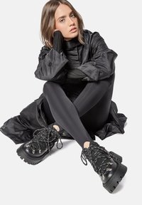 Botas negras hasta el tobillo con suelas gruesas, parte superior texturizada y detalles metálicos en los cordones. Combinadas con mallas negras y un abrigo largo negro.