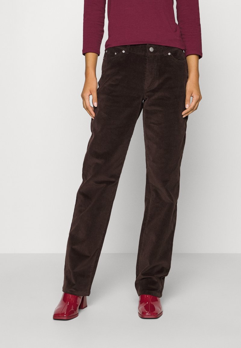 Personne portant un pantalon en velours côtelé marron foncé, un haut à manches longues bordeaux et des bottines rouges brillantes, debout devant un fond blanc uni.
