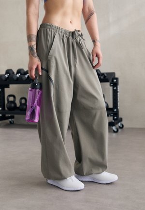 Personne portant un pantalon gris ample et des chaussures blanches tient une bouteille d'eau violette, debout dans une salle de sport avec des kettlebells et des haltères en arrière-plan.