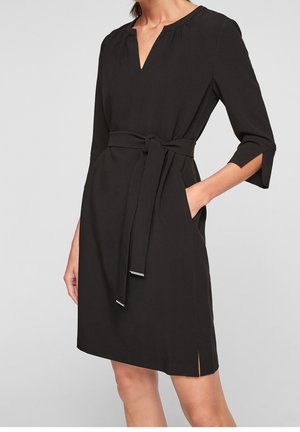 Femme portant une robe noire longueur genou avec des manches trois-quarts, un col en V, des poches latérales et une ceinture nouée à la taille.