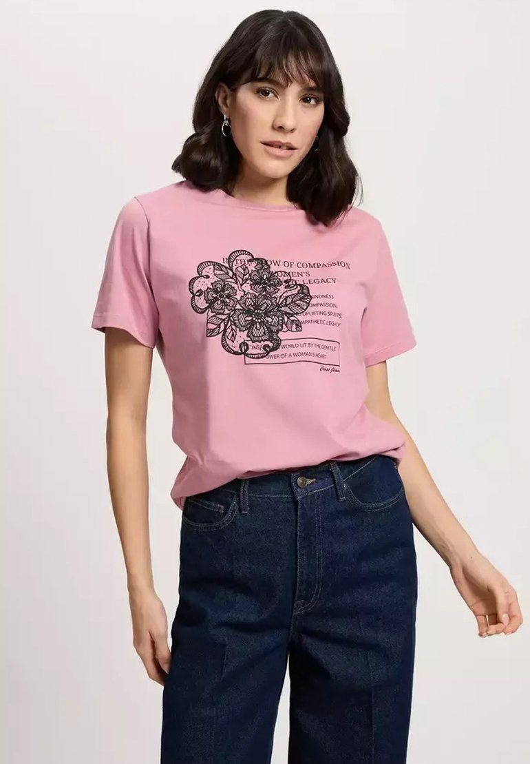 Różowa bawełniana koszulka z czarnym graficznym wzorem kwiatowym i napisem z przodu, okrągły dekolt i krótkie rękawy, zestawiona z wysokim stanem ciemnojeansowymi spodniami.