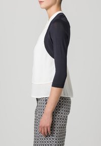 Vit blus med marinblå trekvartsärmar, slät textur, vågiga kanter på ärmarna, i kombination med mönstrade byxor med geometriska former.