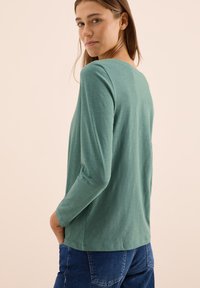Grön långärmad T-shirt med rundad halsringning och mjuk textur, tillsammans med blå jeans. Enkelt mönster och avslappnad passform.