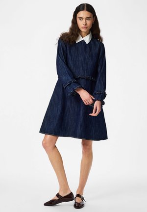 Femme portant une robe longue en denim bleu foncé à manches longues avec un col blanc, des poignets volantés, et des chaussures Mary Jane noires, debout devant un fond blanc.
