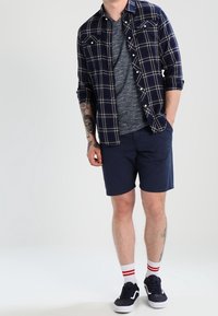 Laivastonsininen ruutupaita harmaan t-paidan päällä, yhdistetty laivastonsinisiin shortseihin. Valkoiset sukat, joissa punaiset raidat, ja mustat lenkkarit täydentävät asun.