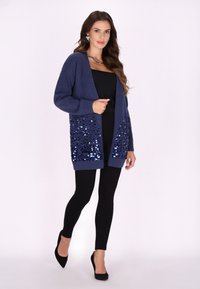 Cardigan navy con apertura frontale, lavorazione a maglia testurizzata e pannelli inferiori decorati con paillettes. Abbinato a leggings neri e tacchi a punta.