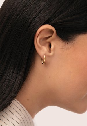 Primer plano de la oreja derecha de una persona con cabello largo y oscuro que lleva un pequeño pendiente de aro dorado en forma de V.