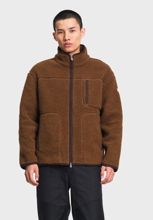 Homme portant une veste polaire marron zippée avec un col montant et des poches poitrine et latérales zippées, debout devant un fond uni.