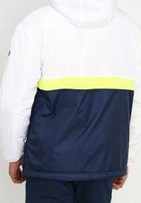 Vit och marinblå jacka med en reflekterande gul rand över ryggen, utrustad med en huva och en jämn, blank yta.