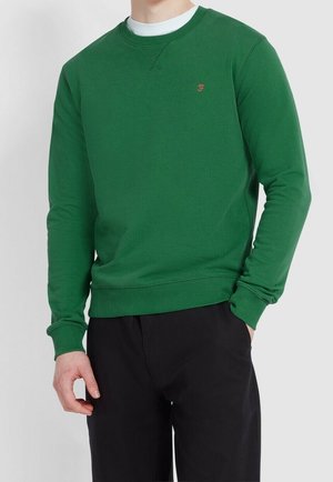 Hombre con sudadera verde de manga larga sobre una camiseta blanca, combinado con pantalones negros, mano en el bolsillo.
